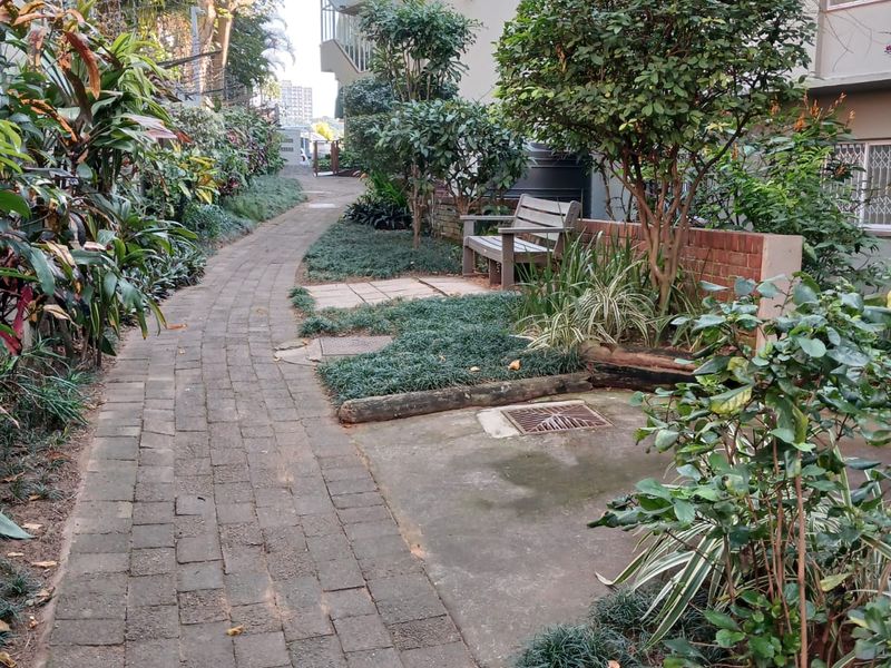 A landscaped garden area featuring a brick paved pathway, mature trees, and a brick retaining wall alongside the retirement village building.