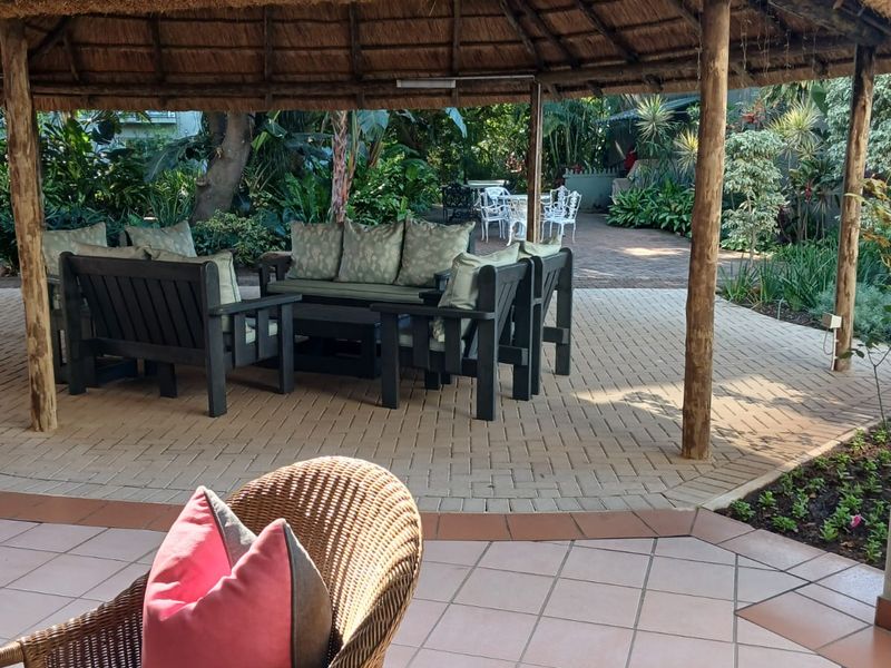 A spacious outdoor patio area featuring a traditional thatched lapa, timber support posts, and a combination of paved and tiled flooring.