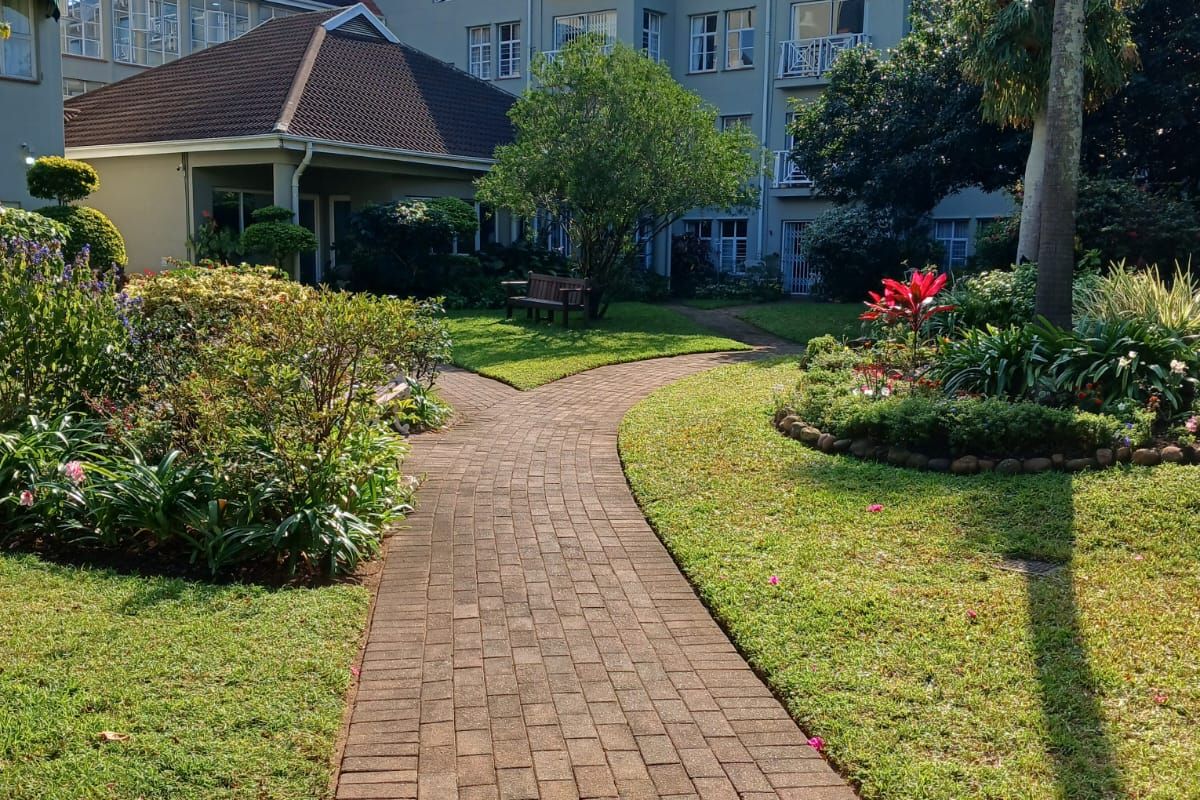 A well-maintained communal garden area featuring a paved walkway leading towards the multi-story retirement village building with balconies and landscaped greenery.