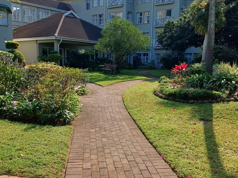 A well-maintained communal garden area featuring a paved walkway leading towards the multi-story retirement village building with balconies and landscaped greenery.
