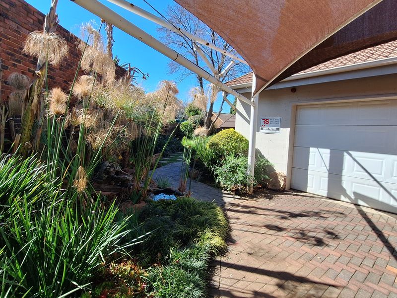Front exterior view featuring a paved driveway, a single garage door, a shade sail structure, and a landscaped garden area with a brick boundary wall.
