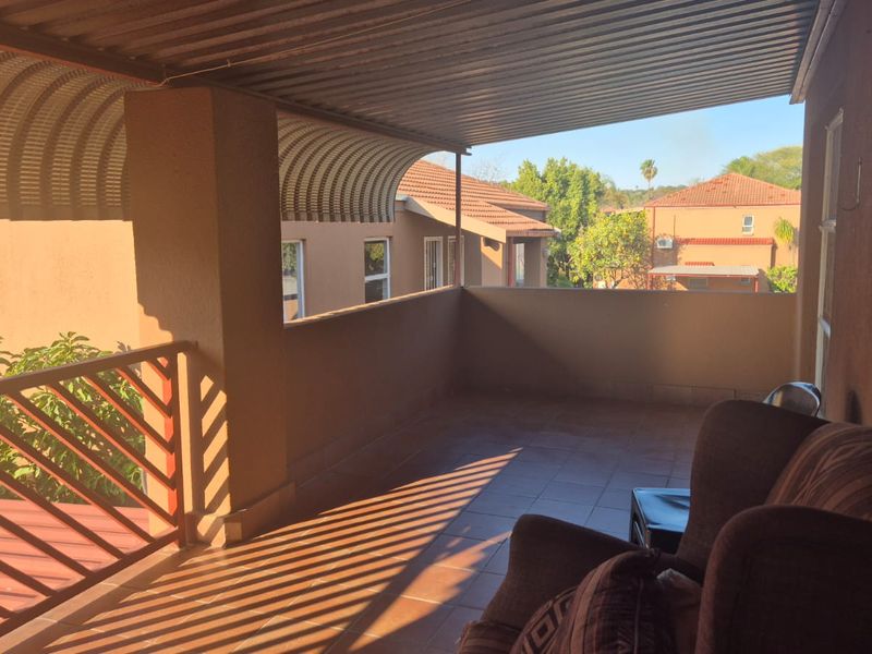A covered balcony featuring tiled flooring, a corrugated metal roof, and a protective railing, offering an outdoor space with views of the surrounding complex.