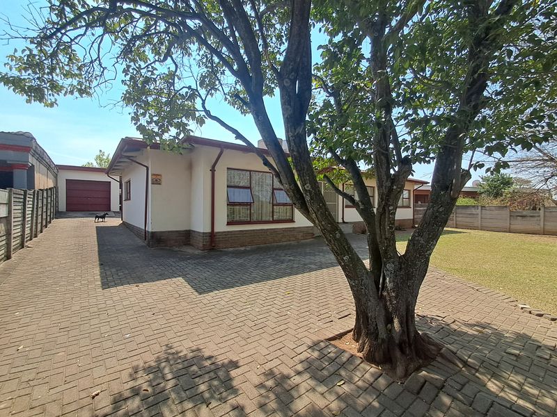 Front view of a single-story house featuring a paved driveway, mature tree, and a detached garage with a brick boundary wall.