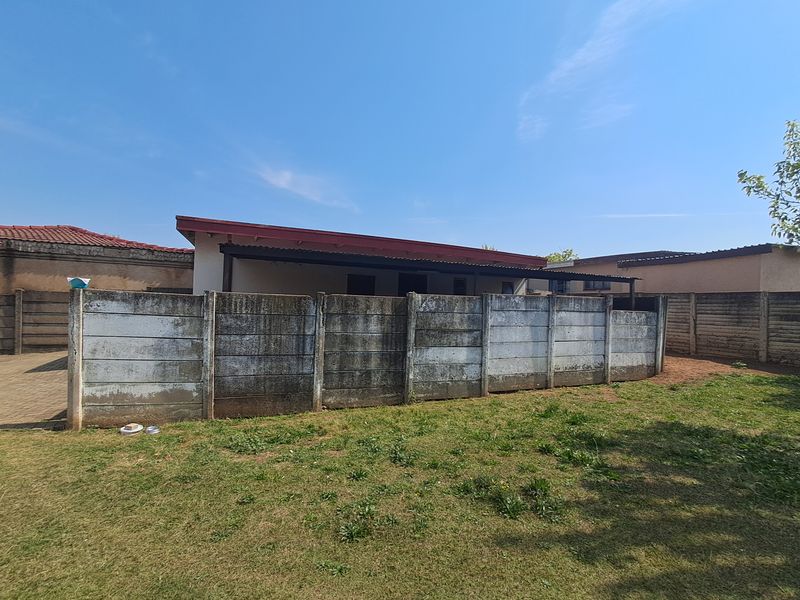 Rear view of the property featuring a covered patio area, a paved driveway, and a precast concrete boundary wall set within a lawned garden.