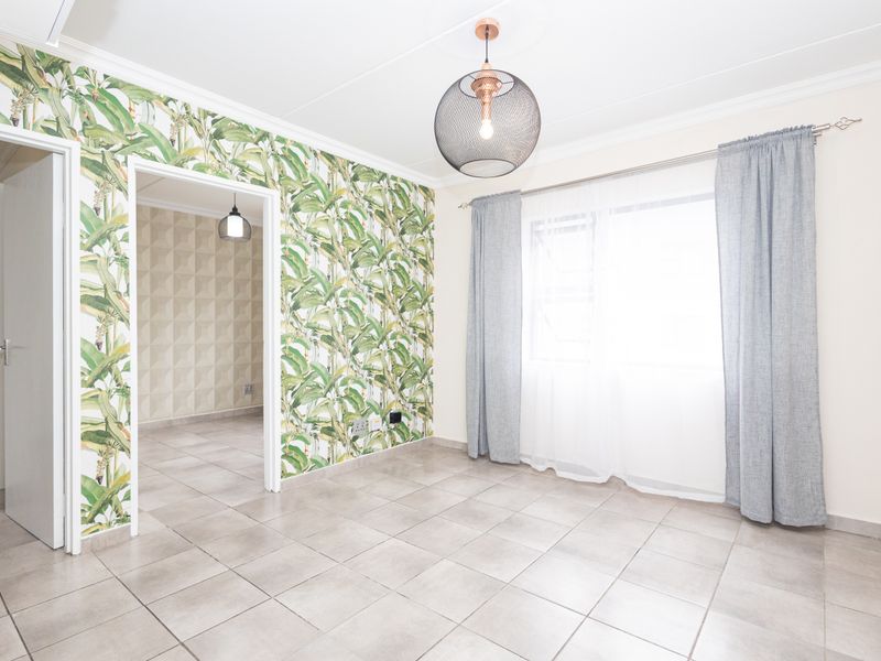 A bright, empty living area featuring neutral tiled flooring, decorative botanical wallpaper, and crown molding, with a view into an adjacent room.