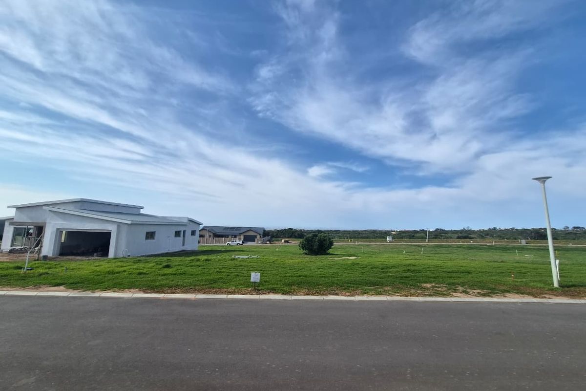 A wide-angle view of a vacant residential plot featuring a paved road, street lighting, and established grass, with neighbouring properties visible in the distance.
