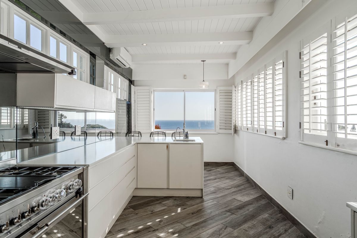 Modern, bright kitchen featuring sleek white cabinetry, a central island, gas stove, and plantation shutters with expansive ocean views.