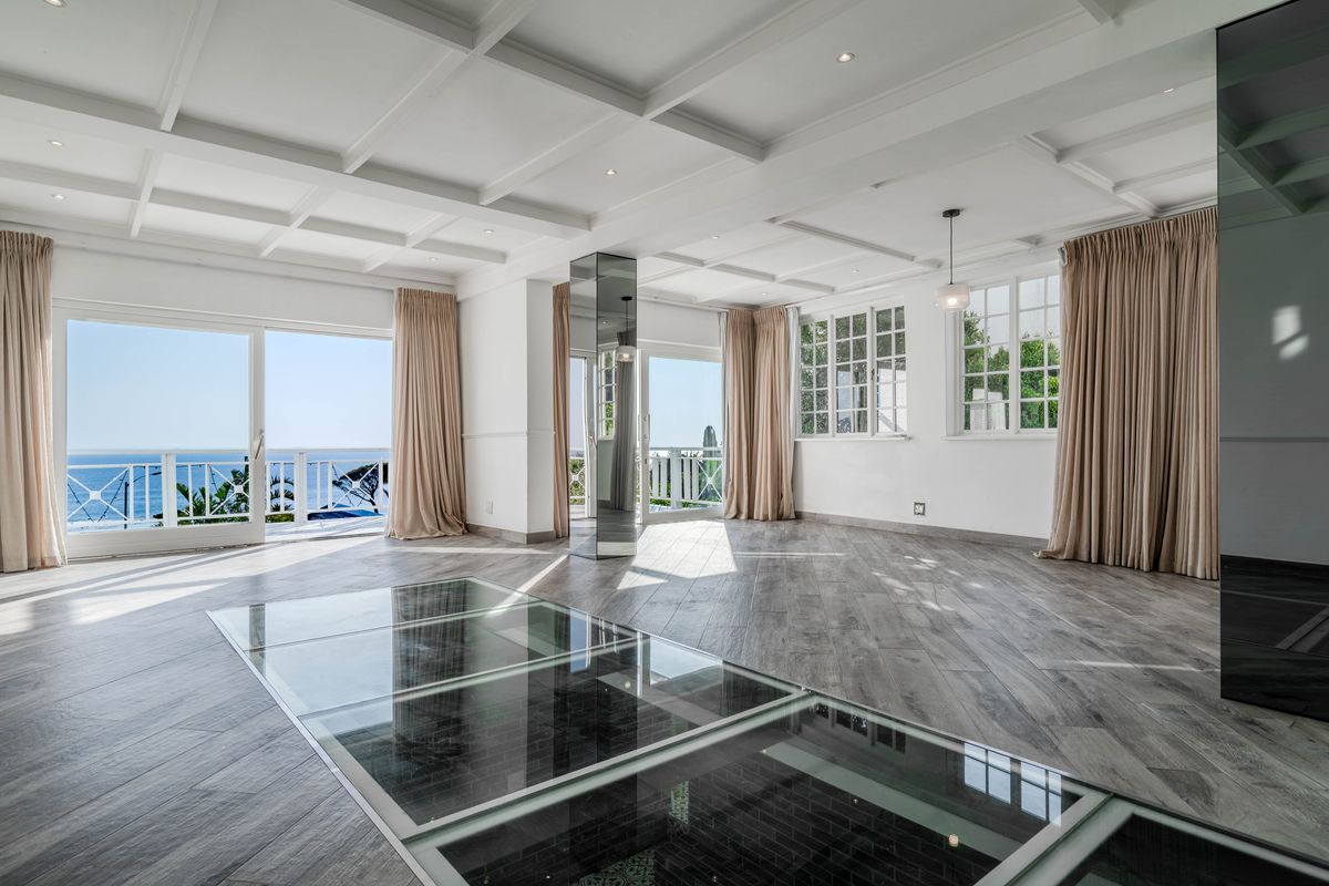 Spacious living area featuring a coffered ceiling, unique glass floor panel, mirrored pillar, and large sliding doors opening to a scenic balcony.