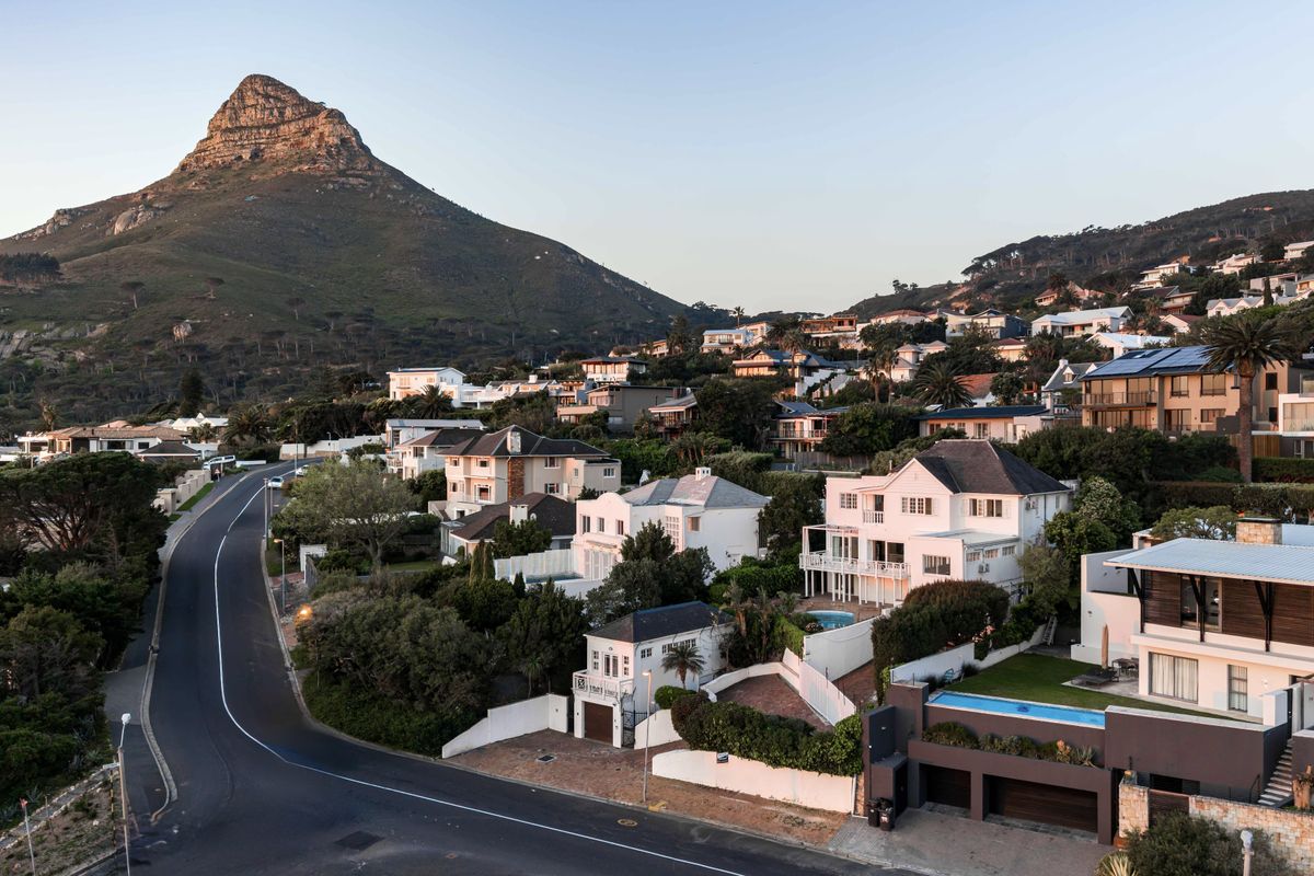 An elevated aerial view of a coastal residential neighborhood featuring multi-story homes, swimming pools, and winding roads set against a prominent mountain backdrop.