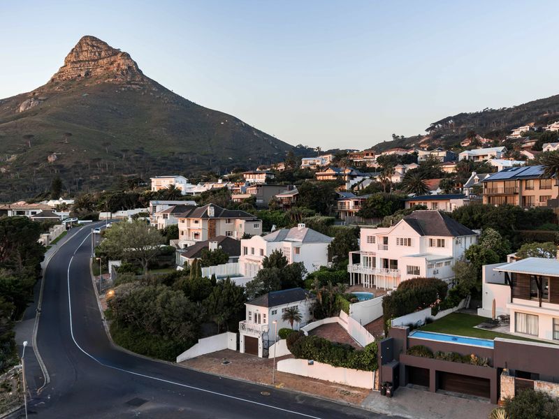 An elevated aerial view of a coastal residential neighborhood featuring multi-story homes, swimming pools, and winding roads set against a prominent mountain backdrop.