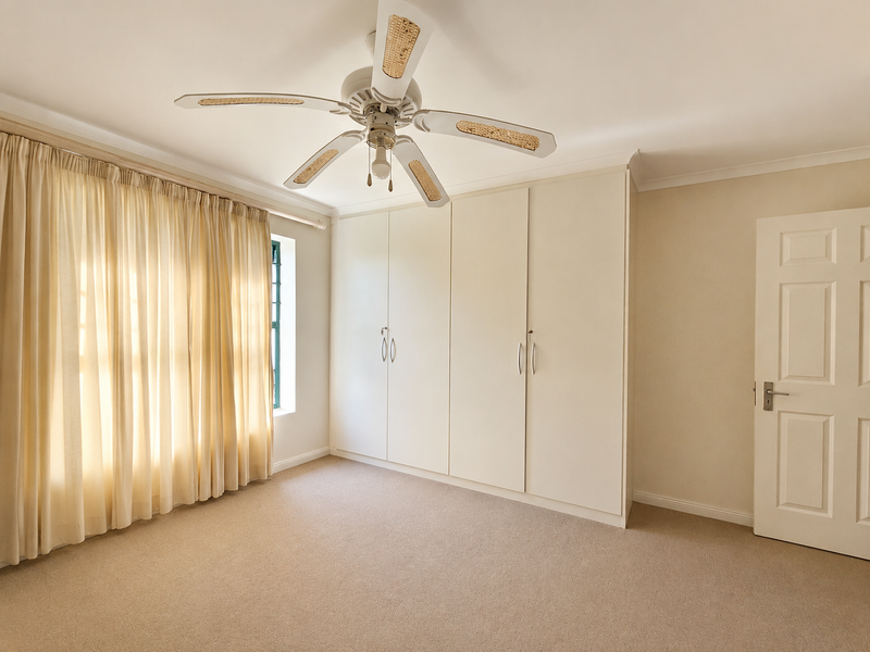 A bright, empty bedroom featuring neutral carpeting, built-in floor-to-ceiling cupboards, and a ceiling fan, offering a clean and versatile space.