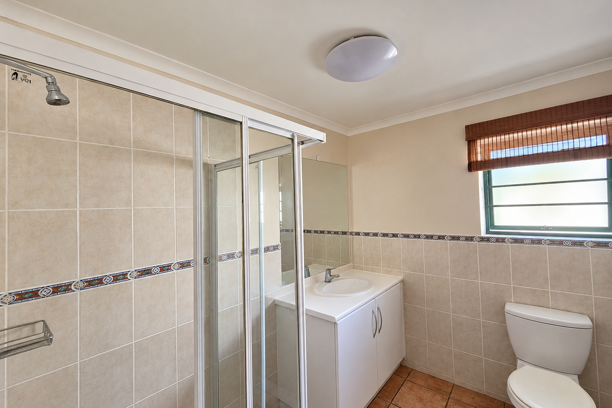 A clean, functional bathroom featuring a shower enclosure, vanity with basin, and toilet, finished with neutral wall tiles and terracotta floor tiles.