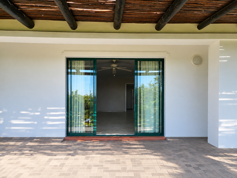 A covered patio area featuring a thatched roof with exposed timber beams, paved flooring, and sliding glass doors leading into the interior.