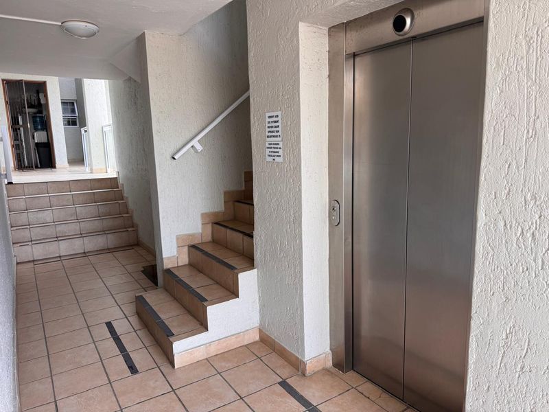 A clean apartment building hallway featuring a stainless steel elevator, tiled flooring, and a staircase with a handrail.