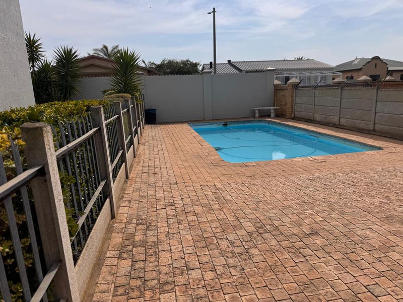 Outdoor pool area featuring a rectangular swimming pool surrounded by paved decking and enclosed by a perimeter wall and metal fencing.