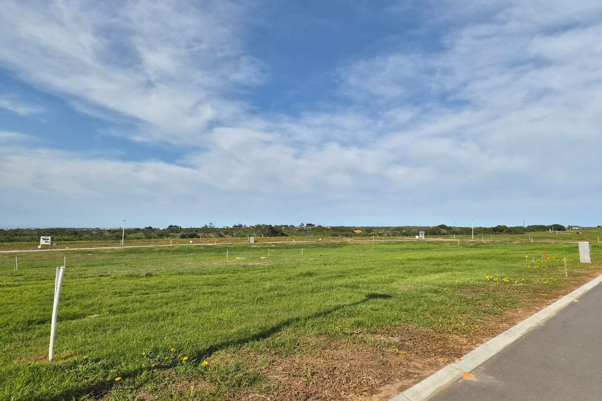 A vacant residential plot featuring level grass terrain, accessible via a paved road with established curbing and boundary markers.