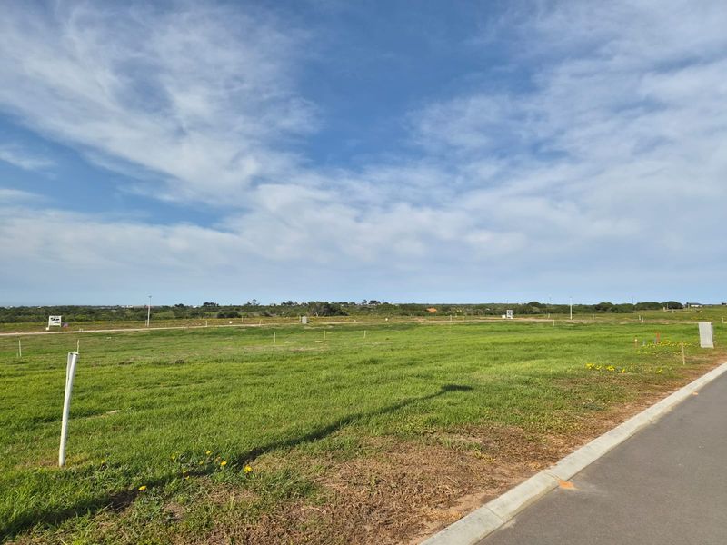 A vacant residential plot featuring level grass terrain, accessible via a paved road with established curbing and boundary markers.