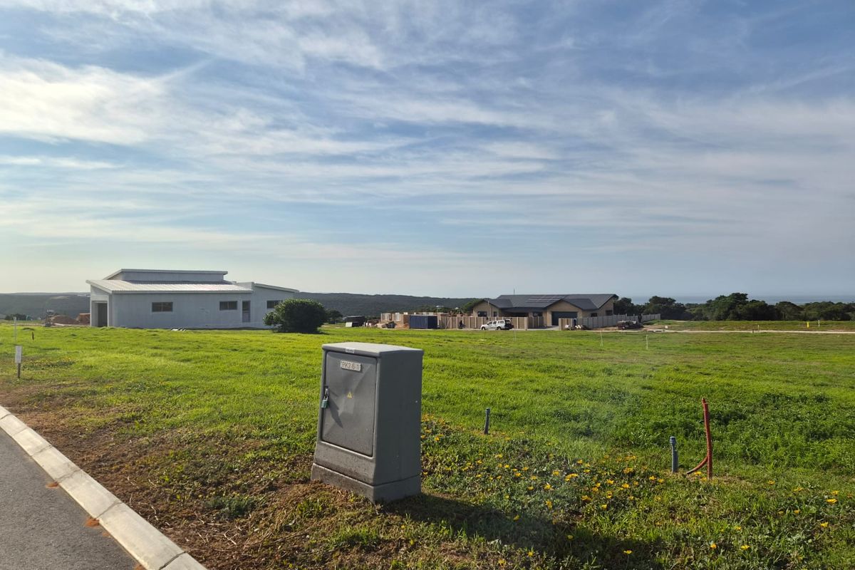A vacant residential plot featuring a grassed landscape, paved curb, and an installed electrical utility box, with neighboring properties visible in the distance.