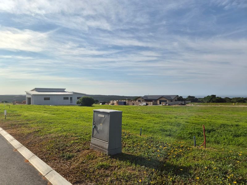 A vacant residential plot featuring a grassed landscape, paved curb, and an installed electrical utility box, with neighboring properties visible in the distance.