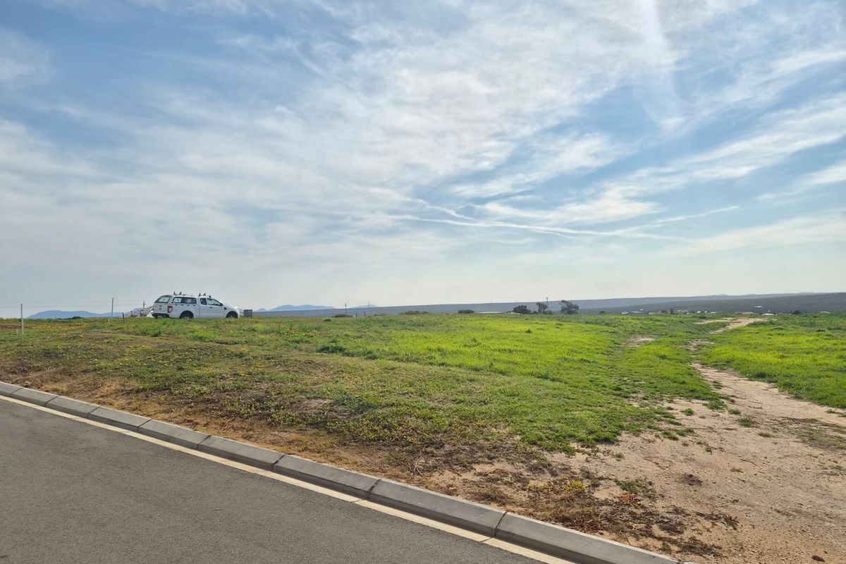 A vacant residential plot featuring a gentle slope, accessible via a paved road with established concrete curbing and perimeter fencing.