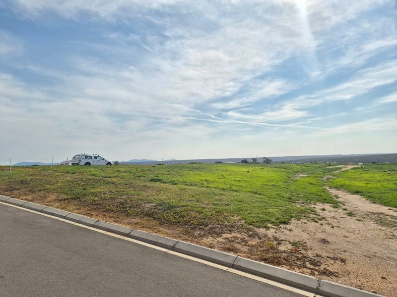 A vacant residential plot featuring a gentle slope, accessible via a paved road with established concrete curbing and perimeter fencing.