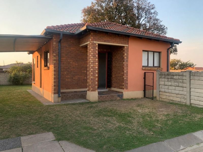 Front view of a detached townhouse featuring a brick and plaster facade, tiled roof, and an attached carport with a surrounding garden area.