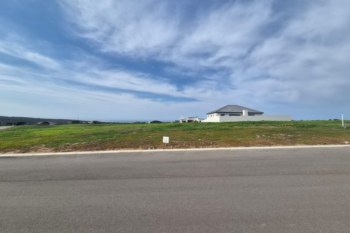 A view of a vacant residential plot from the street, featuring open grassed land with surrounding neighborhood development.
