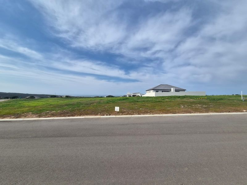 A view of a vacant residential plot from the street, featuring open grassed land with surrounding neighborhood development.