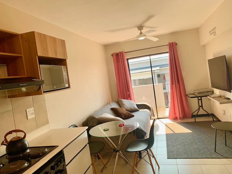 Open-plan living area featuring a compact kitchen with built-in cabinetry, electric hob, and extractor fan, leading to a balcony through sliding glass doors.