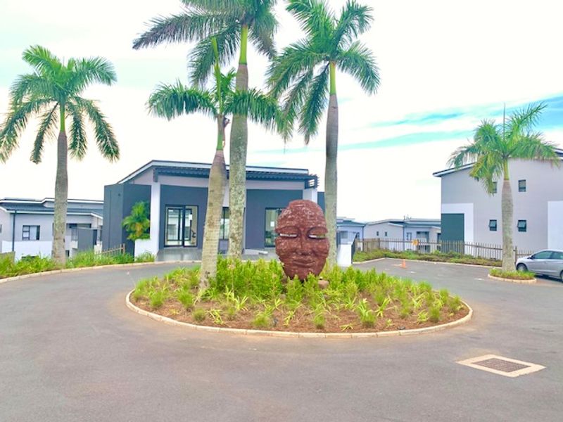 A well-maintained residential complex entrance featuring a landscaped roundabout with palm trees and modern architectural building facades.