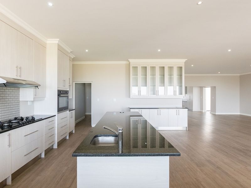 Spacious open-plan kitchen featuring modern light-toned cabinetry, stone countertops, a central island with a sink, and integrated appliances in a bright, neutral setting.
