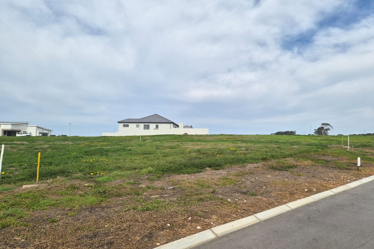 A vacant residential plot featuring established grass cover, adjacent to a paved road with kerbing, offering a clear building site.