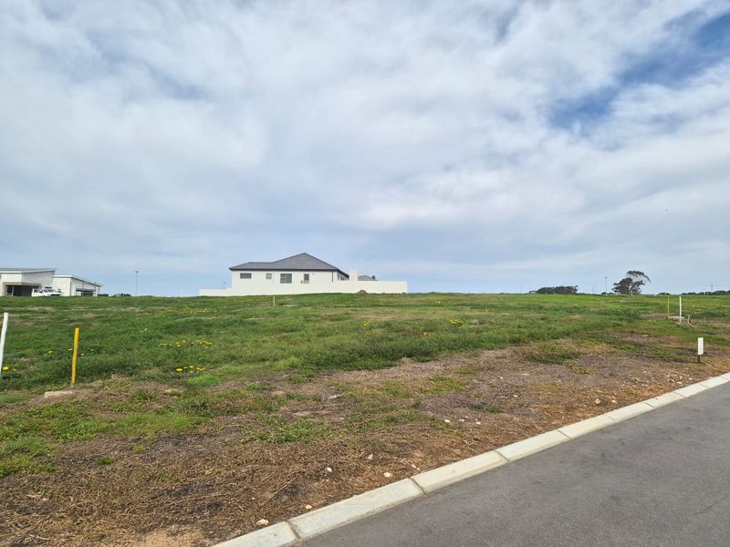 A vacant residential plot featuring established grass cover, adjacent to a paved road with kerbing, offering a clear building site.