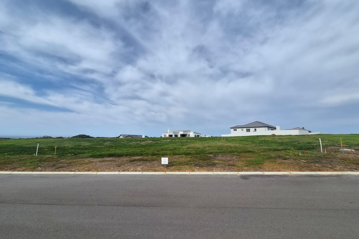 A vacant residential plot of land featuring a grassed surface, situated along a paved road with surrounding neighborhood development visible in the distance.