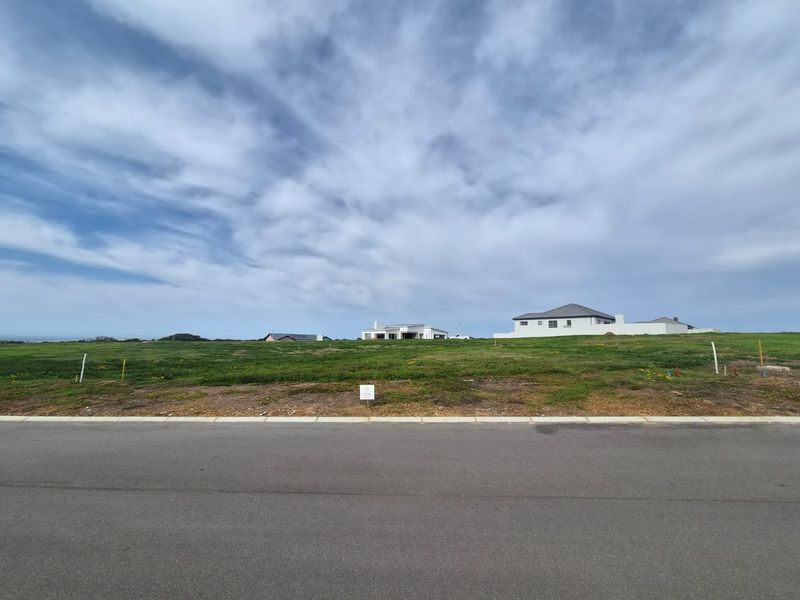 A vacant residential plot of land featuring a grassed surface, situated along a paved road with surrounding neighborhood development visible in the distance.