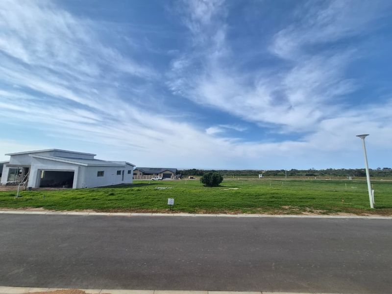 A wide-angle view of a vacant residential plot featuring a paved road, street lighting, and established grass, with neighbouring properties visible in the distance.