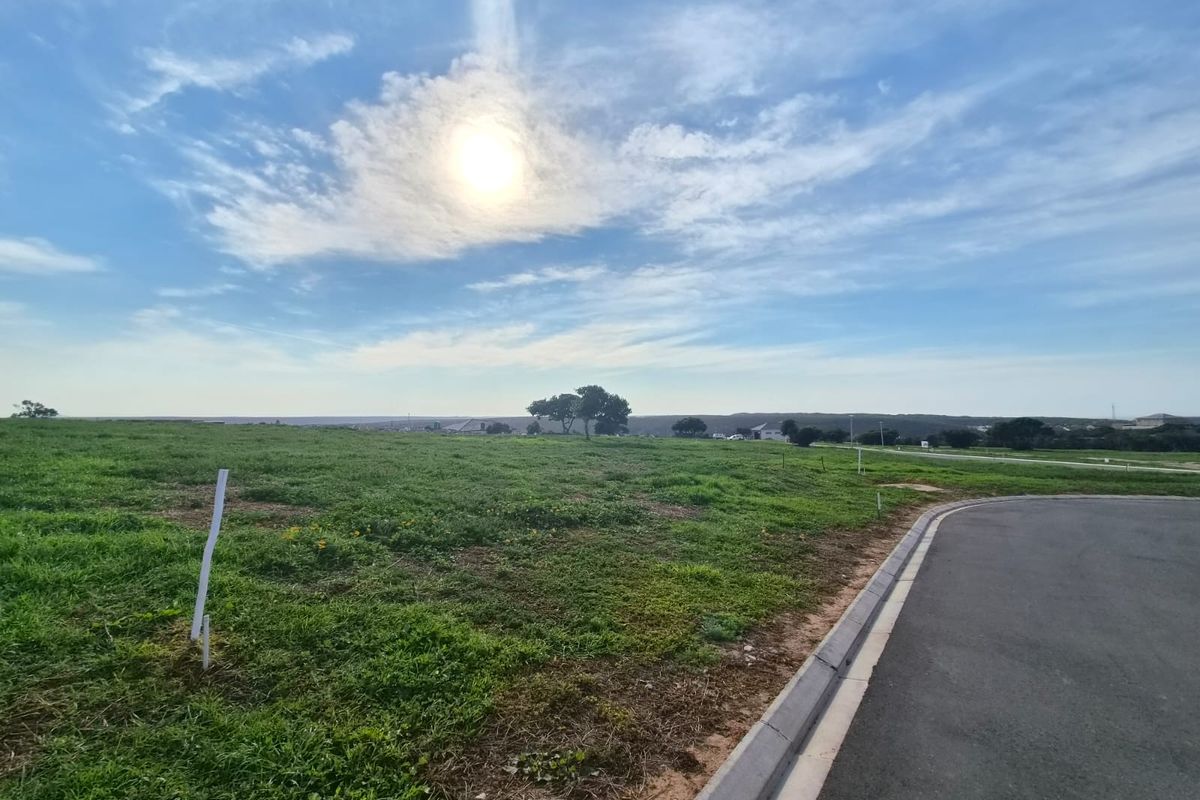 A vacant residential plot featuring a paved road frontage, concrete curbing, and open grassy terrain with clear views of the surrounding landscape.