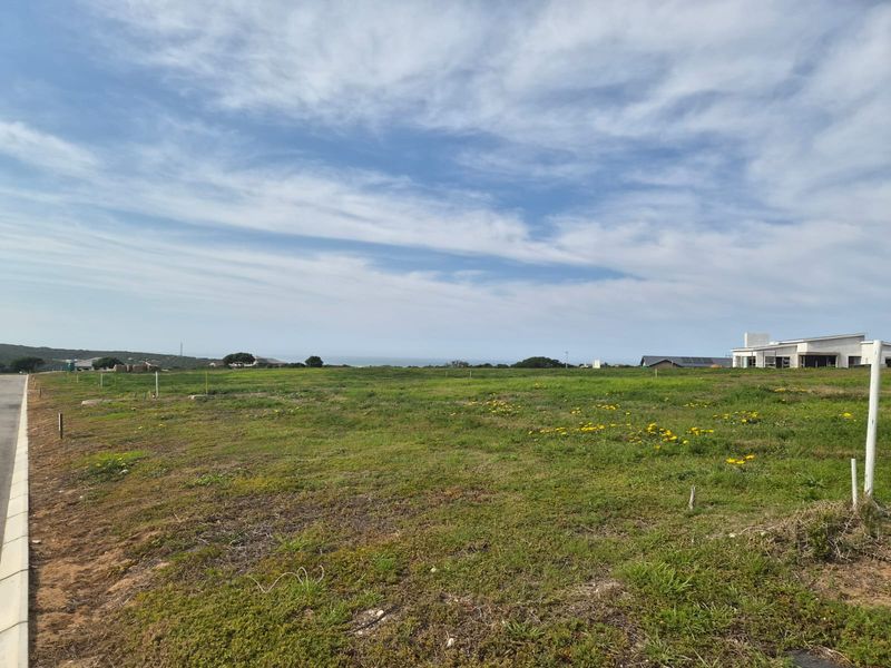 A vacant residential plot featuring a level grassy landscape with distant ocean views and adjacent modern housing developments.