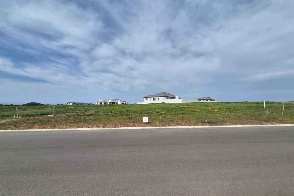 A vacant residential plot of land featuring a grassed surface, situated along a paved road with surrounding neighborhood development visible in the background.