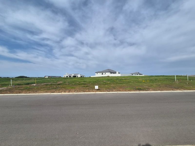 A vacant residential plot of land featuring a grassed surface, situated along a paved road with surrounding neighborhood development visible in the background.