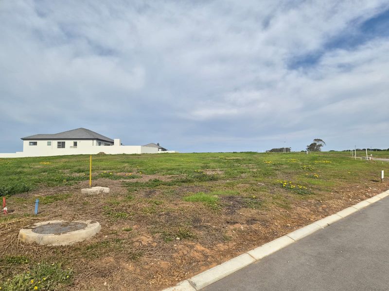 A vacant residential plot featuring a gentle slope, established road access, and visible utility connections, ready for development in a quiet neighborhood.