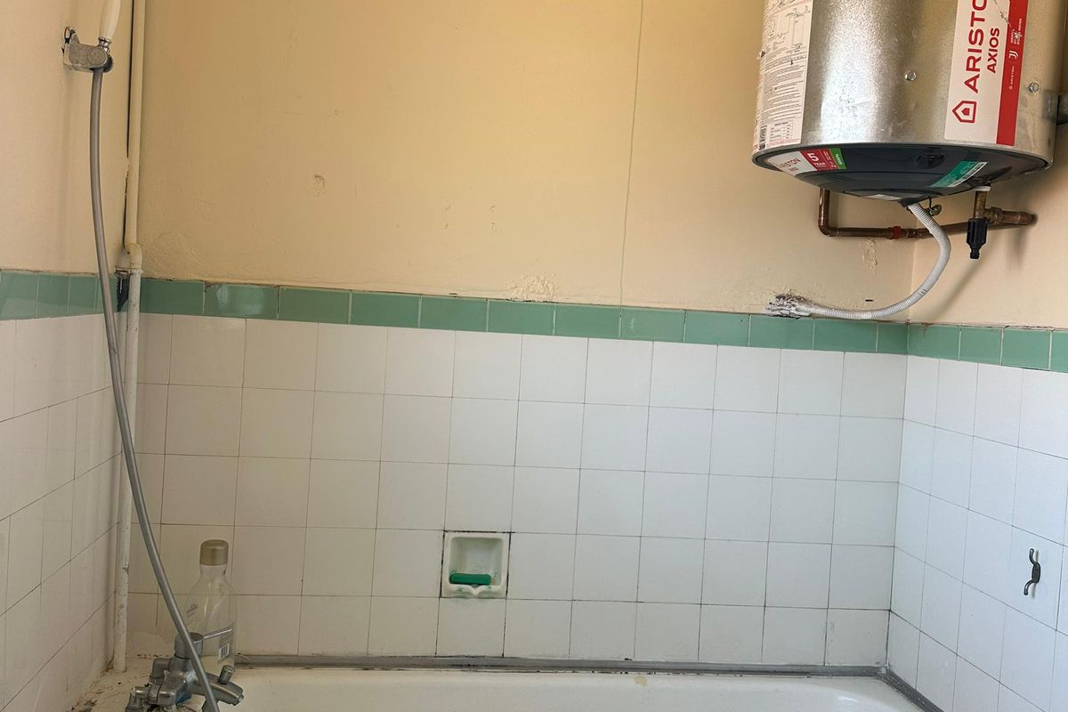 A dated bathroom featuring a bathtub, wall-mounted shower mixer, and an exposed electric water heater with visible plumbing.