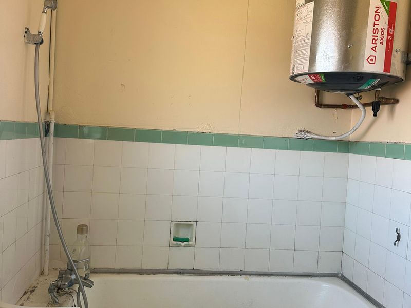A dated bathroom featuring a bathtub, wall-mounted shower mixer, and an exposed electric water heater with visible plumbing.