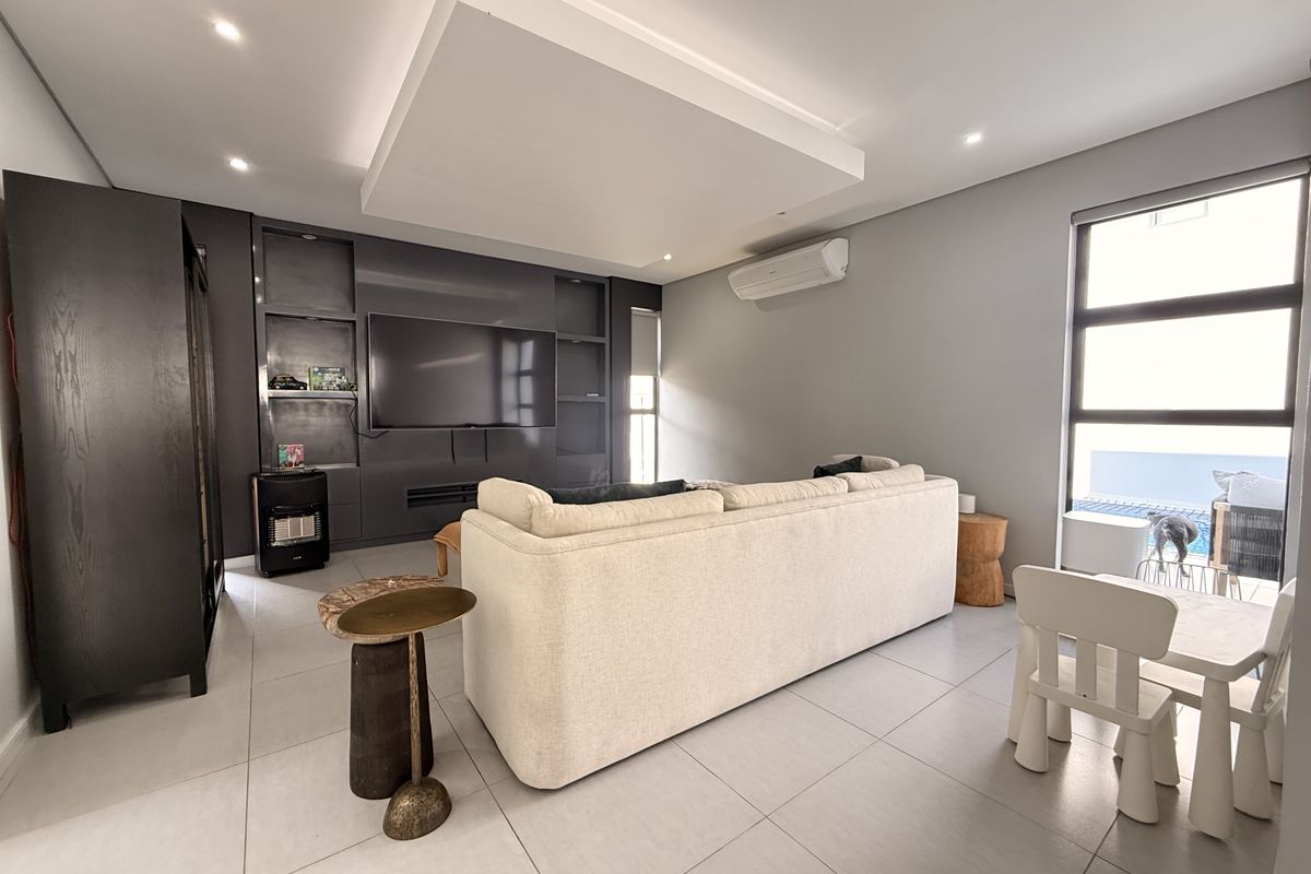 A modern living room featuring a large built-in media unit, recessed ceiling detail, split-system air conditioning, and large windows overlooking the pool area.