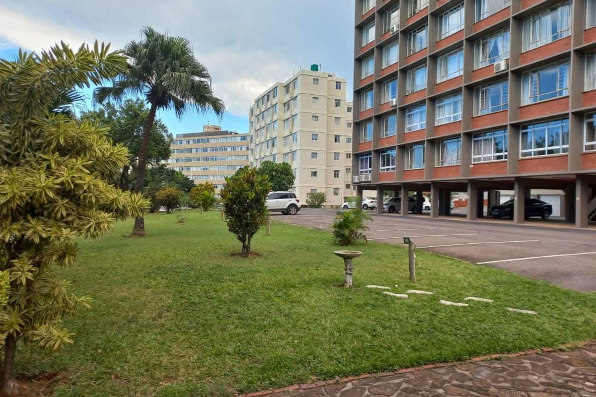Exterior view of a multi-story apartment complex featuring a landscaped garden area and a paved parking lot at the base of the building.