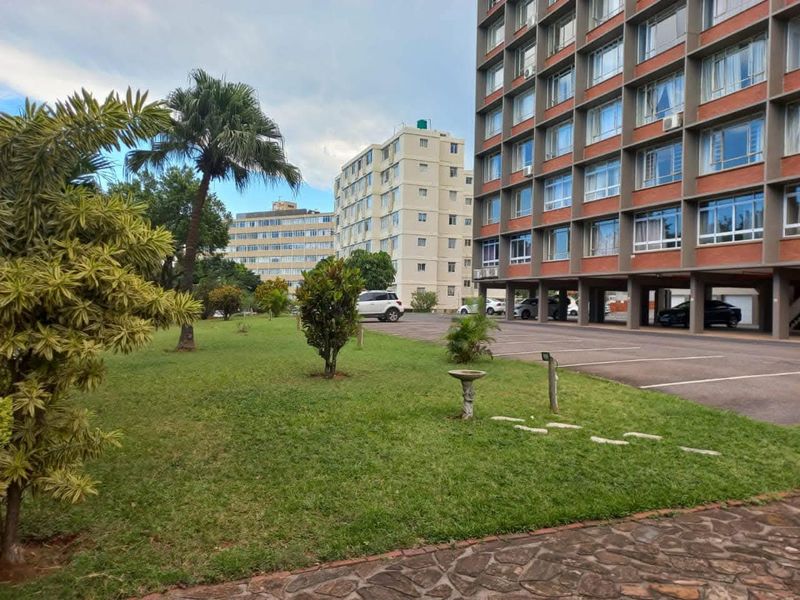 Exterior view of a multi-story apartment complex featuring a landscaped garden area and a paved parking lot at the base of the building.