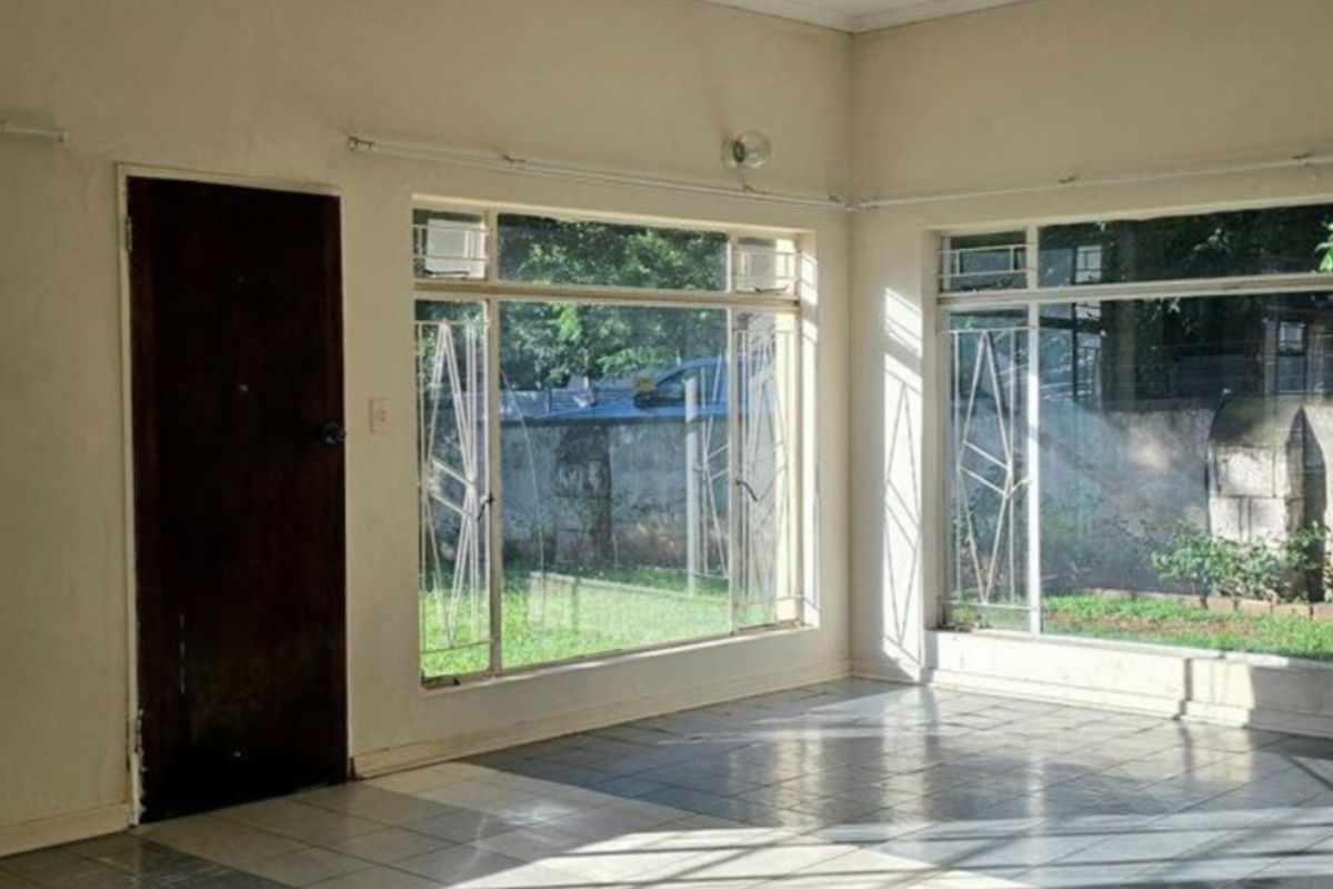 An empty living room featuring tiled flooring, large windows with security bars, and a timber door, offering a bright space with garden views.