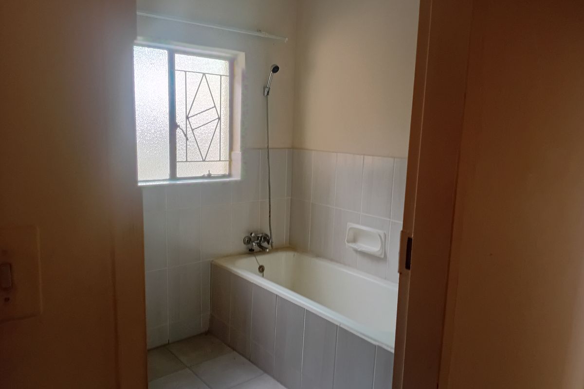 A functional bathroom featuring a built-in bathtub with shower mixer, tiled walls, and a window with security bars.