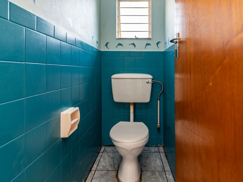 A compact bathroom featuring blue wall tiling, a standard toilet, and a louver window, requiring cosmetic updates to modernize the space.