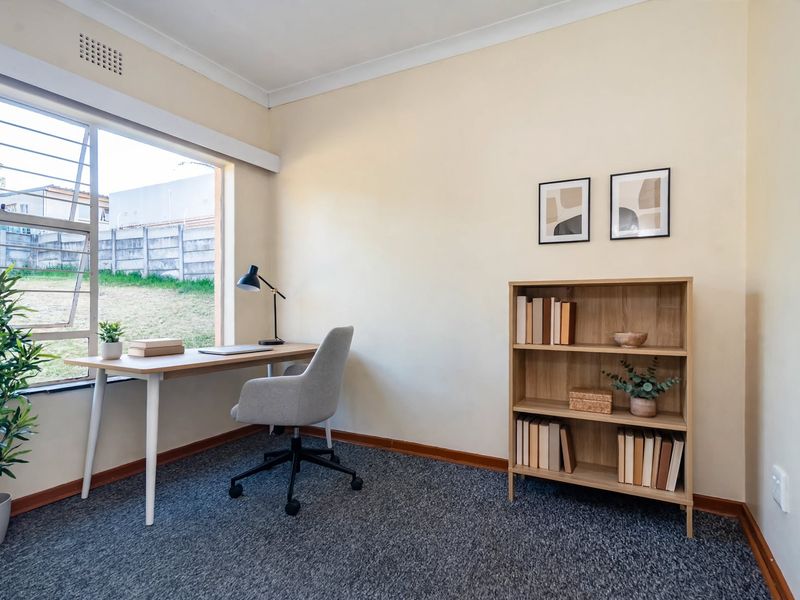 A bright home office featuring carpeted flooring, large window with security bars, and neutral wall finishes, providing a functional workspace.